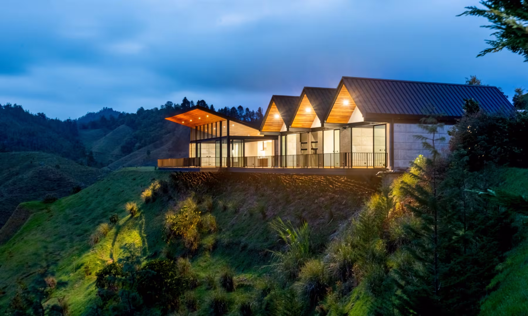 Fachada iluminada al anochecer con cubiertas a dos aguas y deck sobre la ladera, casa campestre en El Retiro