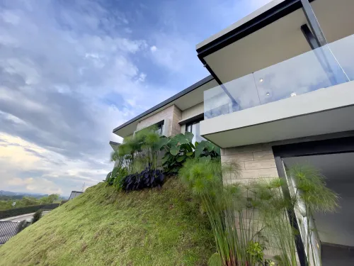 Fachada lateral de casa campestre moderna en venta con balcon de vidrio y jardin en ladera.